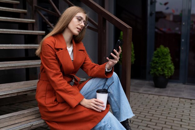 Una joven elegante se sienta en las escaleras junto a un café popular con una taza de café y mira un