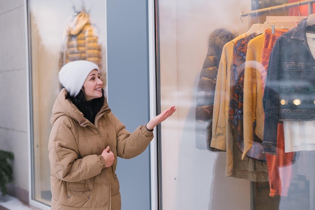 Una joven elegante con sombrero y abrigo en la ciudad mira hacia las ventanas de las tiendas de ropa