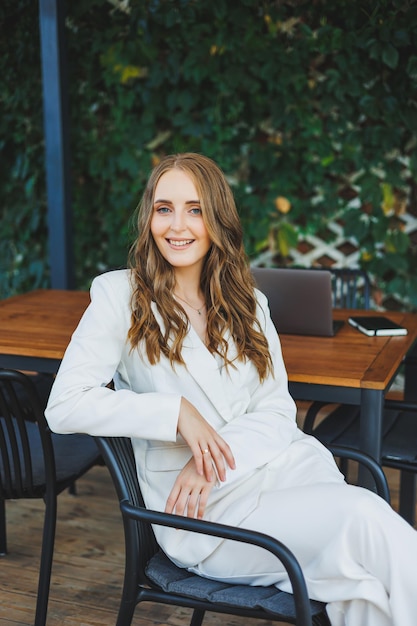 Una joven elegante en un traje clásico blanco con una sonrisa en su rostro está de pie en la terraza de una cafetería de verano