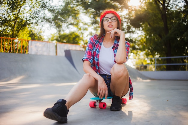 Joven elegante vestida con ropa juvenil se sienta en una patineta en un skatepark. Verano, día soleado y brillante