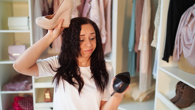 Joven eligiendo zapatos de tacón alto en el armario Mujer aturdida de cabello negro con camiseta blanca casual mirando a la cámara con la boca abierta mientras sostiene diferentes zapatos de tacón alto en las manos en el armario