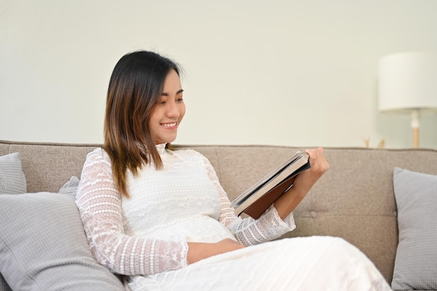 Una joven embarazada asiática relajada leyendo un libro mientras descansa en su sala de estar