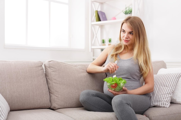 Joven embarazada comiendo ensalada verde. Rubia expectante irreconocible sentada en el sofá y con bocadillos frescos. Concepto de nutrición y embarazo saludable