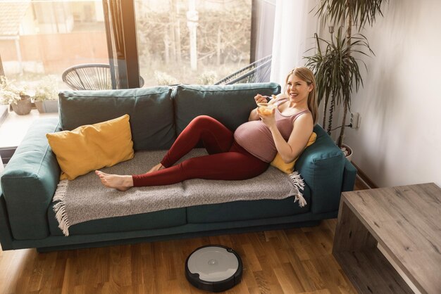 Joven embarazada relajándose en el sofá de casa por la mañana mientras la aspiradora robot está trabajando y limpiando la sala de estar.