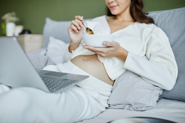 Joven embarazada sentada en la cama con una laptop de rodillas y viendo algo durante su desayuno en el dormitorio