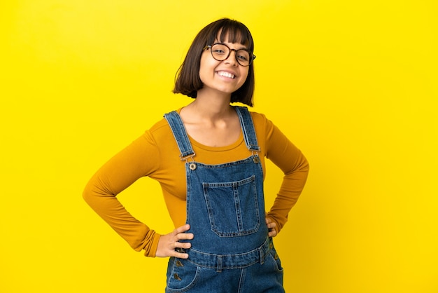 Joven embarazada sobre fondo amarillo aislado posando con los brazos en la cadera y sonriendo