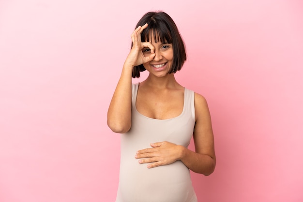Joven embarazada sobre fondo rosa aislado que muestra el signo de ok con los dedos