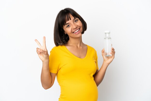 Joven embarazada sosteniendo una botella de agua aislado sobre fondo blanco sonriendo y mostrando el signo de la victoria