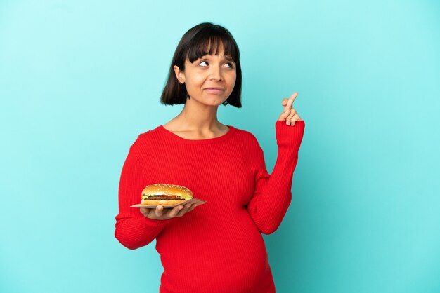 Joven embarazada sosteniendo una hamburguesa sobre antecedentes aislados con los dedos cruzando y deseando lo mejor