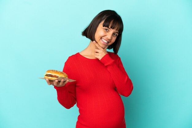 Joven embarazada sosteniendo una hamburguesa sobre antecedentes aislados feliz y sonriente