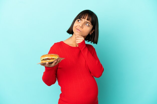 Joven embarazada sosteniendo una hamburguesa sobre antecedentes aislados y mirando hacia arriba