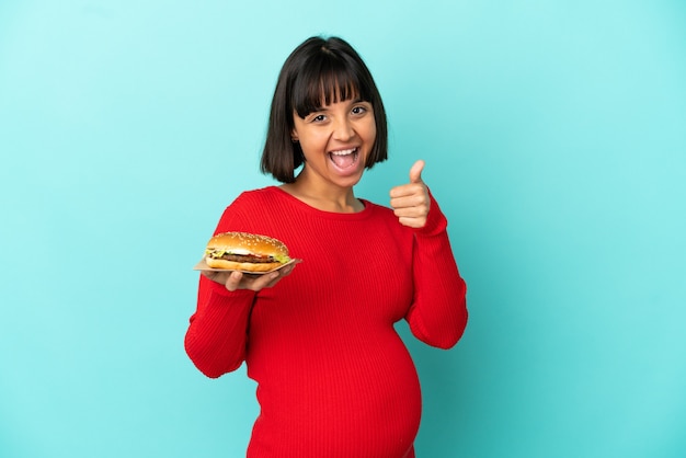 Joven embarazada sosteniendo una hamburguesa sobre antecedentes aislados con los pulgares hacia arriba porque algo bueno ha sucedido