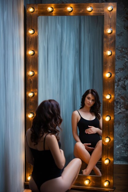 Una joven embarazada en un traje negro se sienta cerca de un hermoso espejo de madera con lámparas.