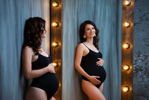 Una joven embarazada en un traje negro se sienta cerca de un hermoso espejo de madera con lámparas.