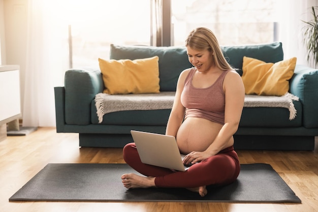 Una joven embarazada usando una computadora portátil mientras se relaja en su sala de estar por la mañana.