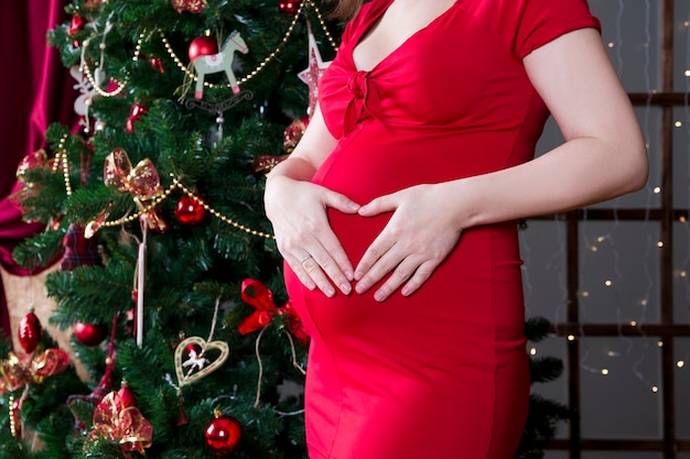 Joven embarazada en vestido rojo posando en el fondo del concierto de vacaciones de árbol de Navidad