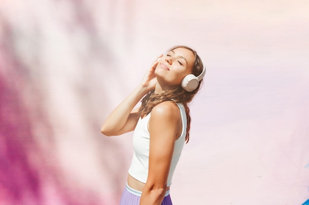Una joven emocionada con una blusa y una falda con auriculares escuchando música caminando bailando con las manos en alto al aire libre contra una pared rosa
