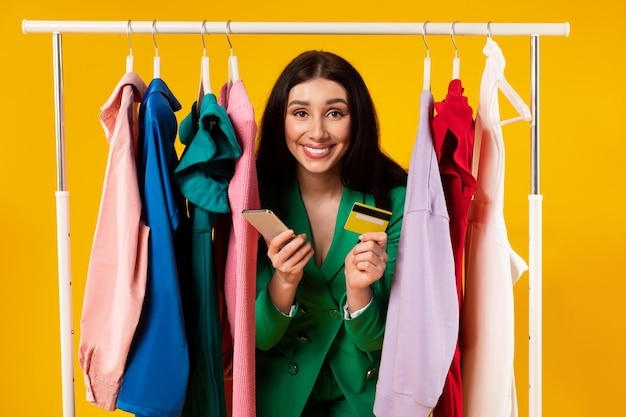 Una joven y emocionada clienta sosteniendo un teléfono celular y una tarjeta de crédito posando cerca de un riel de ropa sobre un fondo amarillo