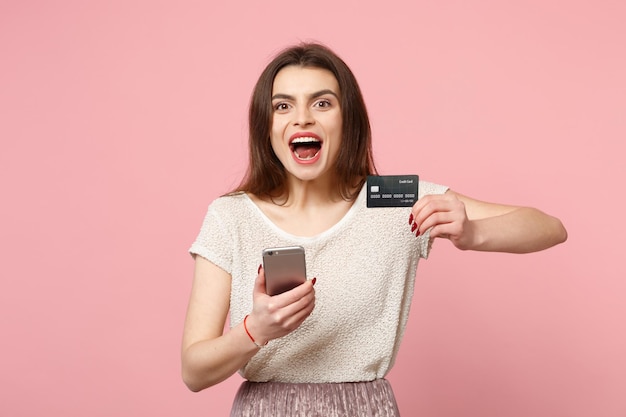 Una joven emocionada con ropa ligera informal posando aislada en un fondo rosa pastel. Concepto de estilo de vida de las personas. Simulacros de espacio de copia. Usar teléfono móvil, escribir mensajes sms, tener tarjeta bancaria de crédito.