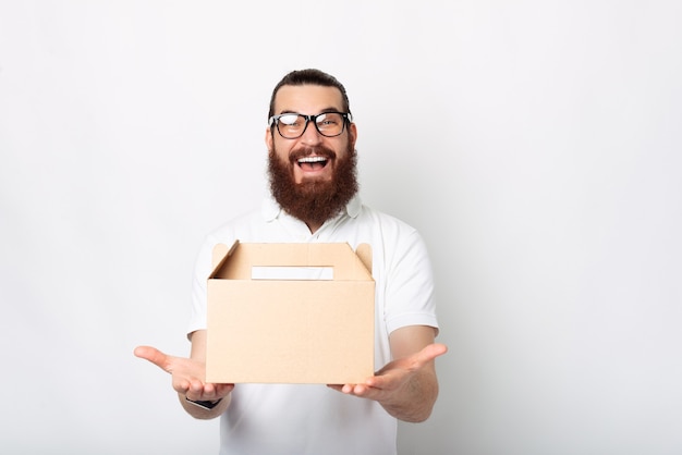 Un joven emocionado con una caja de entrega está sonriendo asombrosamente a la cámara cerca de la pared blanca