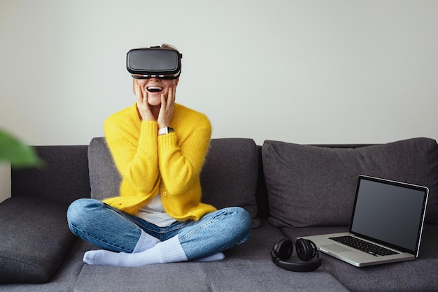 Joven emocionado sentado en el sofá y usando auriculares de realidad virtual en casa