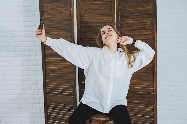 Una joven emocional con una camisa blanca de algodón gesticula y mira la pantalla de un aparato moderno Una chica con un teléfono móvil se toma una selfie