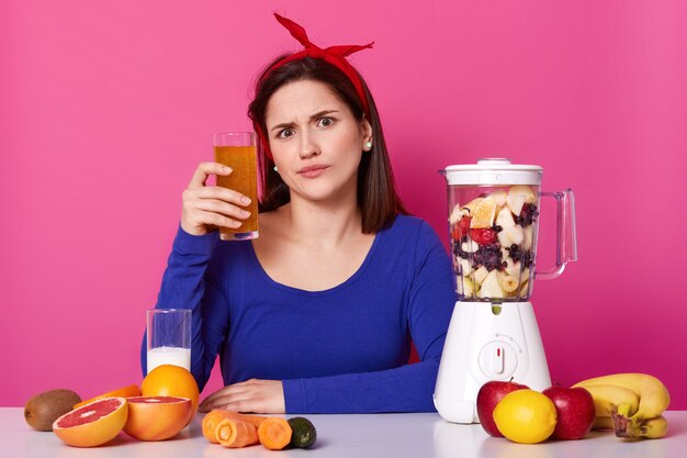 Joven emocional europea se sienta a la mesa con equipo de cocina, sosteniendo un vaso de batido de naranja