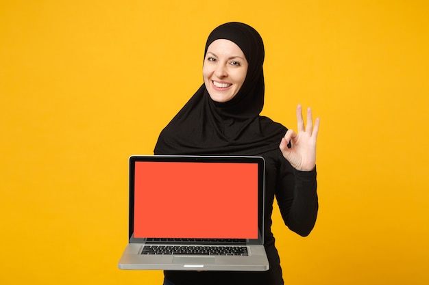 Joven empleada musulmana árabe en hijab ropa negra mantenga y trabaje computadora portátil aislada en retrato de pared amarilla. Concepto de estilo de vida religioso de la gente.