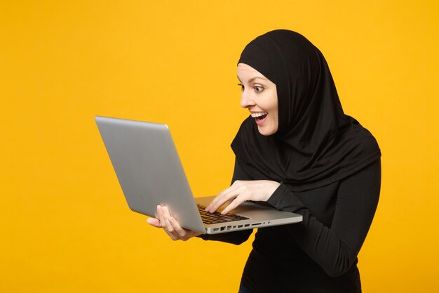 Joven empleada musulmana árabe en hijab ropa negra mantenga y trabaje computadora portátil aislada en retrato de pared amarilla. Concepto de estilo de vida religioso de la gente.