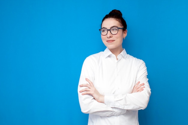 Una joven empleada de oficina con camisa blanca y gafas se para con los brazos cruzados sobre un fondo azul