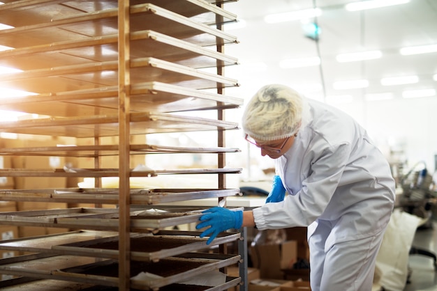 Foto joven empleada de panadería trabajadora en paños estériles empujando el estante con hojalata llena de galletas recién horneadas.