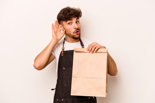 Joven empleado hispano sosteniendo una bolsa para llevar aislada de fondo blanco tratando de escuchar un chisme