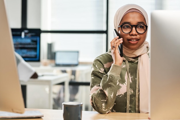 Joven empleado musulmán confiado en hijab y anteojos consultando a un cliente en el teléfono móvil mientras se sienta