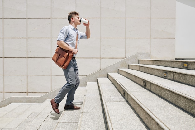 Joven emprendedor soñoliento subiendo las escaleras cuando se apresura a trabajar y bebiendo café para llevar