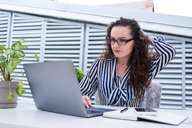 Joven emprendedora exitosa en ropa casual está sonriendo y mirando una pantalla de computadora, trabajando con una computadora portátil en una terraza de un café al aire libre