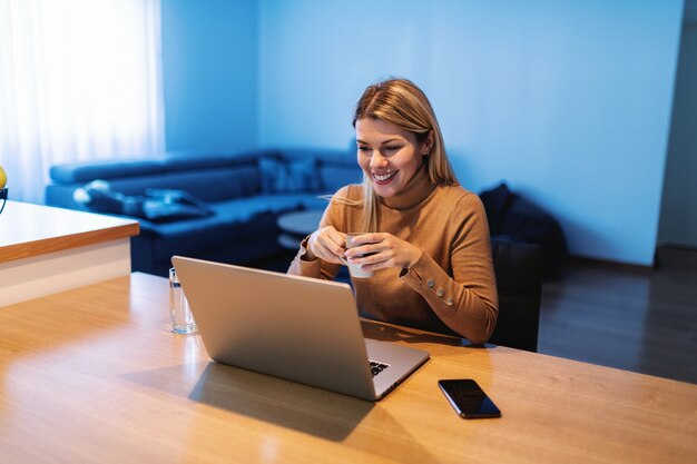 Joven emprendedora rubia impresionante y dedicada sentada en su casa, tomando café y trabajando en un proyecto importante durante el brote de covid 19.