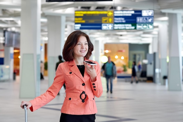 Joven empresaria en el aeropuerto con equipaje graba un mensaje de audio por teléfono y sonríe