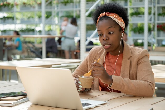 Joven empresaria africana sentada en la mesa frente a la computadora portátil y trabajando durante el almuerzo de negocios en la cafetería