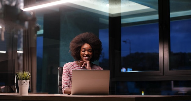 Joven empresaria afroamericana independiente que trabaja con una computadora portátil en el espacio de coworking nocturno