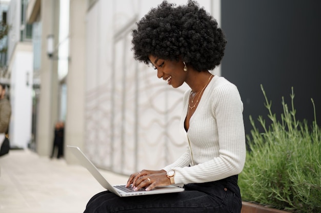 Joven empresaria afroamericana trabajando con una laptop sentada en el banco de la ciudad