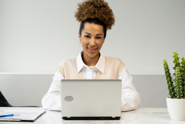Joven empresaria afroamericana utiliza una computadora portátil en el lugar de trabajo y mira a la cámara