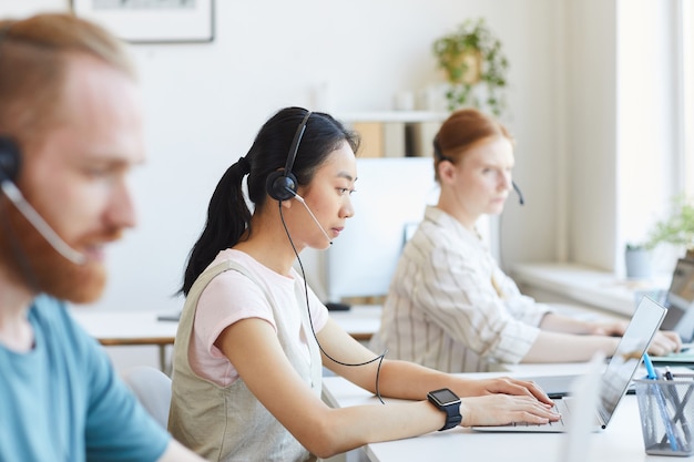 Joven empresaria asiática en auriculares escribiendo en la computadora portátil en su lugar de trabajo con otros colegas que trabajan en la oficina