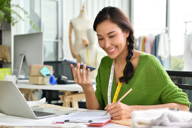 Joven empresaria asiática / diseñadora de moda que trabaja en el estudio y por teléfono con el cliente