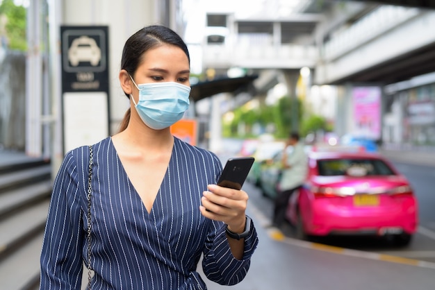 Joven empresaria asiática con máscara con teléfono en la estación de taxis en las calles de la ciudad