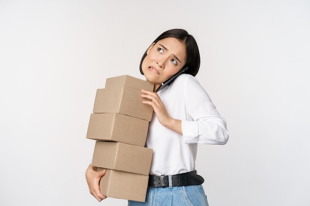 Una joven empresaria asiática responde una llamada telefónica hablando por teléfono móvil mientras lleva un montón de cajas con pedidos sobre fondo blanco.