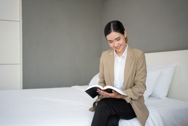 Joven empresaria asiática sentada en la cama leyendo un libro en la habitación del hotel durante un viaje de negocios