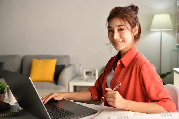 Joven empresaria asiática sentada en casa trabajando en una computadora portátil. Mujer asiática que estudia en línea usando información de navegación portátil o comprando en una tienda de Internet