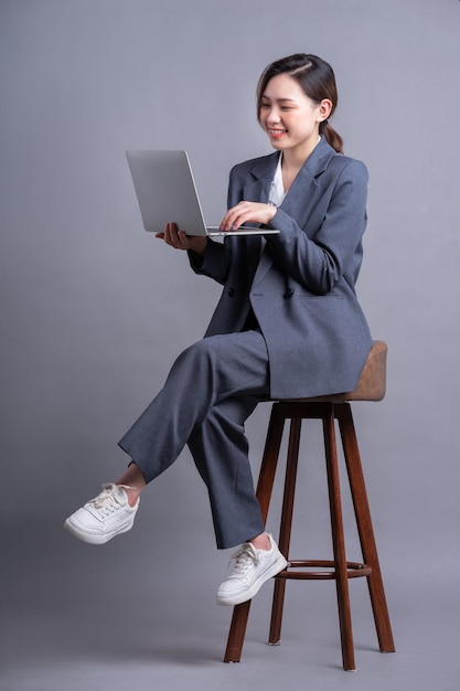 Joven empresaria asiática sentada en una silla y usando una laptop con fondo gris