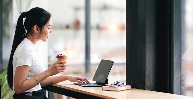 Joven empresaria asiática sosteniendo una taza y usando una tableta digital mientras está sentada en un espacio de trabajo