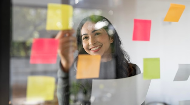 Joven empresaria atractiva y sonriente que usa notas adhesivas en una pared de vidrio para escribir un plan de negocios de estrategia para que el desarrollo crezca hacia el éxito